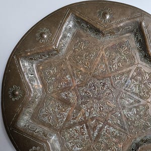 May include: A round, metallic, bronze-toned plate featuring an intricate, embossed eight-pointed star design at its center. The surface is decorated with detailed floral patterns and textured filigree, with small flower motifs along the rim.