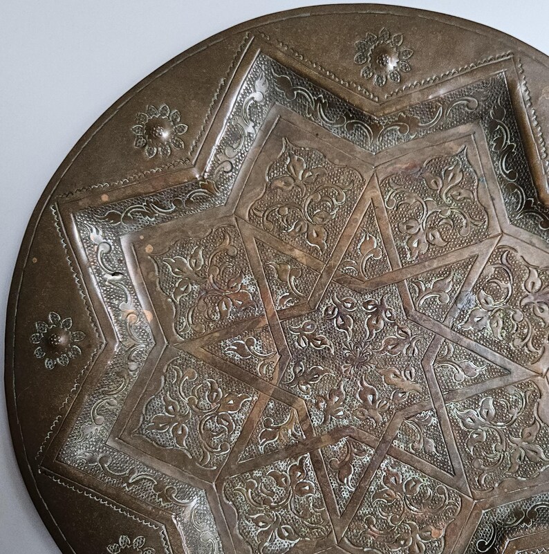 May include: A round, metallic, bronze-toned plate featuring an intricate, embossed eight-pointed star design at its center. The surface is decorated with detailed floral patterns and textured filigree, with small flower motifs along the rim.