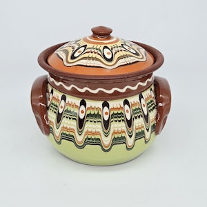 May include: A round ceramic pot with a matching lid and two side handles. The base is light green, featuring a decorative pattern of vertical, wavy lines in brown, cream, and dark green. The lid and rim are brown with a similar wavy design.