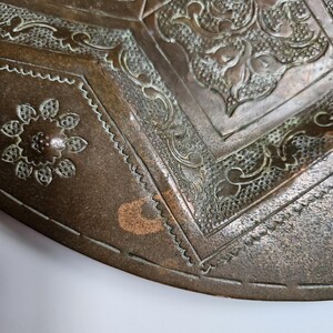 May include: A circular bronze tray featuring intricate embossed patterns, including a central floral motif surrounded by geometric borders and stippled textures. The metal surface displays a weathered patina with visible oxidation.