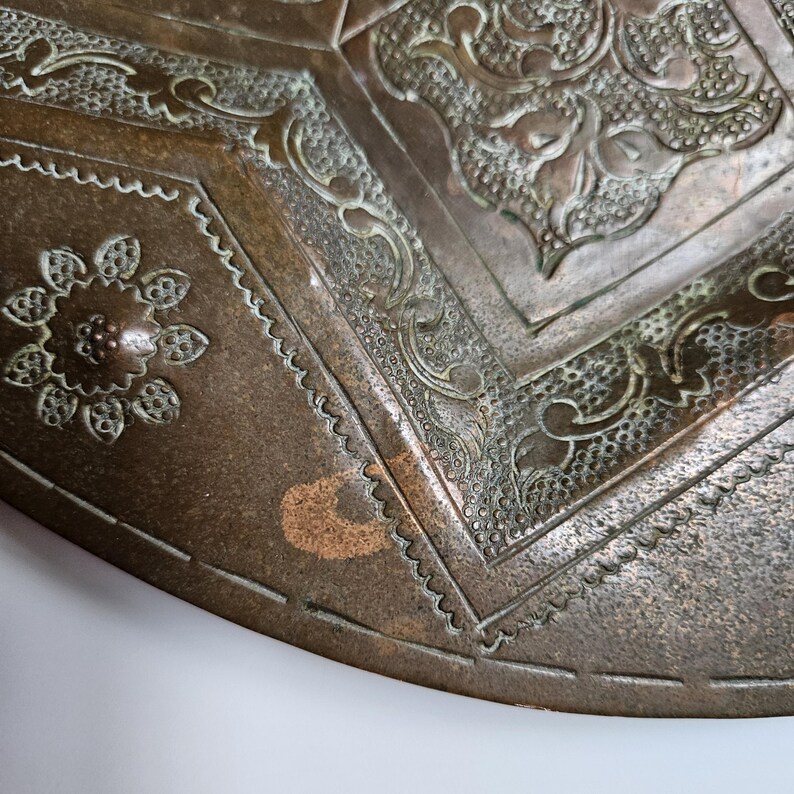 May include: A circular bronze tray featuring intricate embossed patterns, including a central floral motif surrounded by geometric borders and stippled textures. The metal surface displays a weathered patina with visible oxidation.