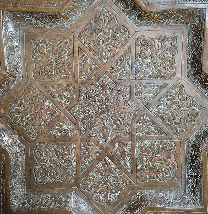 May include: A square copper tray featuring an intricate, embossed eight-pointed star design at its center. The surface is decorated with detailed floral motifs and textured patterns, showcasing a warm, metallic brown finish throughout.