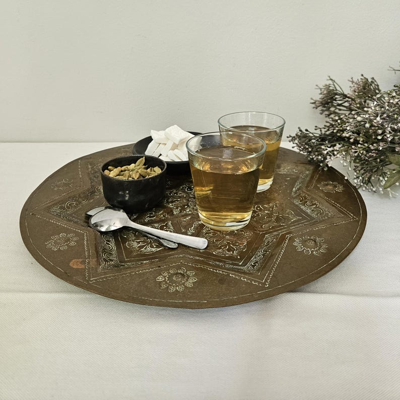 May include: A round, etched brass tray featuring a central star motif and floral patterns. It holds two clear glass tumblers filled with amber liquid, a small black bowl of green cardamom pods, a small dish of white sugar cubes, and a silver spoon.