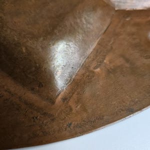 May include: A close-up view of a hammered copper bowl featuring a warm, metallic brown tone with visible surface texture and subtle indentations. The curved edge of the bowl shows a polished finish with natural variations in the metal color.