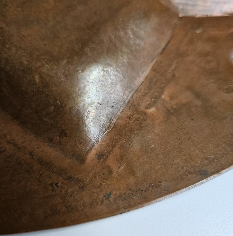 May include: A close-up view of a hammered copper bowl featuring a warm, metallic brown tone with visible surface texture and subtle indentations. The curved edge of the bowl shows a polished finish with natural variations in the metal color.