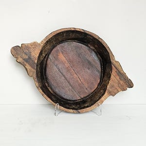 May include: A rustic, wooden bowl with a dark interior and a natural, weathered exterior. The bowl has a round, shallow interior and two decorative, wing-like extensions on either side. The wood grain is visible throughout the bowl.