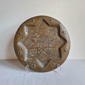 May include: A round brass decorative plate featuring an intricate eight-pointed star design in the center. The surface is etched with detailed floral patterns and small circular motifs along the outer rim, showcasing a weathered metallic finish.