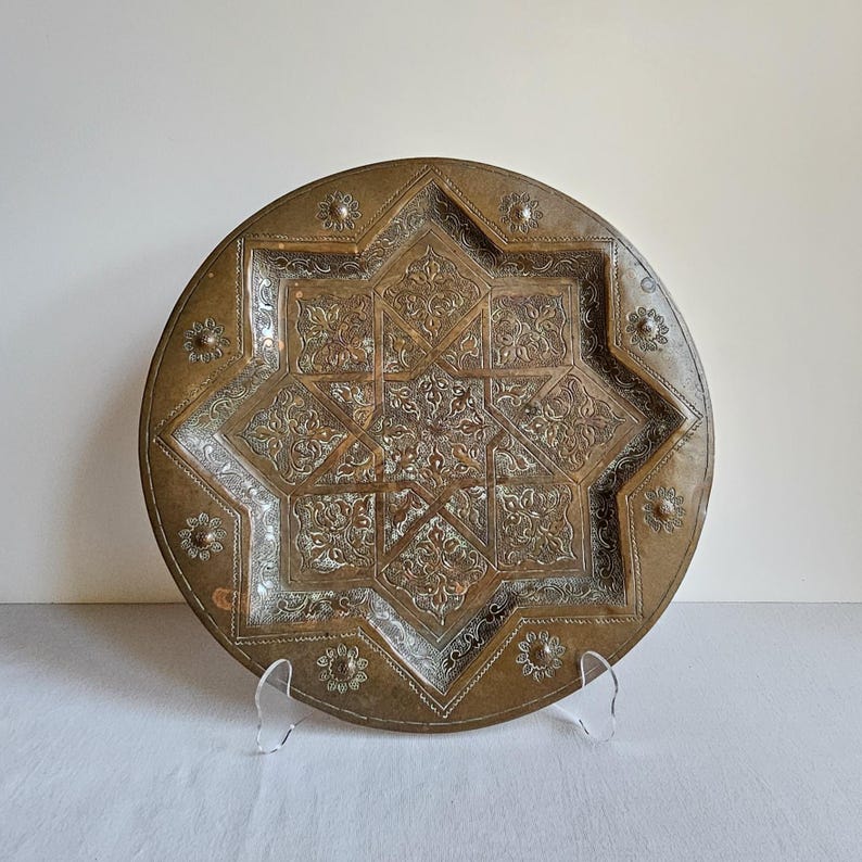 May include: A round brass decorative plate featuring an intricate eight-pointed star design in the center. The surface is etched with detailed floral patterns and small circular motifs along the outer rim, showcasing a weathered metallic finish.