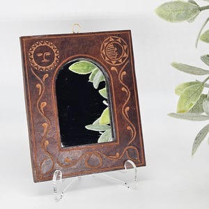 May include: A small, rectangular brown leather mirror with an arched opening. The frame is decorated with carved sun and leaf designs, and a swirling vine pattern. The mirror is displayed on a clear acrylic stand.