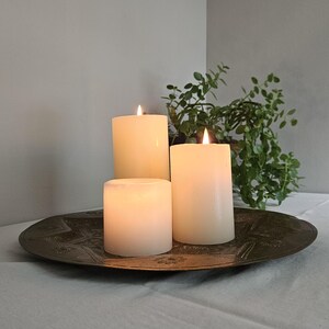 May include: Three cylindrical white wax candles of varying heights are lit and arranged on a round, dark metallic tray featuring an embossed star pattern. The candles emit a soft, warm glow, creating a contrast against the dark metal surface.
