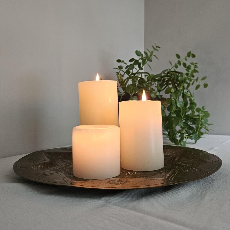 May include: Three cylindrical white wax candles of varying heights are lit and arranged on a round, dark metallic tray featuring an embossed star pattern. The candles emit a soft, warm glow, creating a contrast against the dark metal surface.