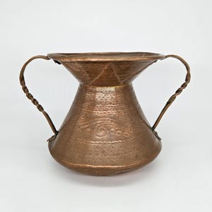 May include: A hammered copper vase with two twisted handles. The vase has a wide, flared rim and a bulbous body with decorative patterns. The copper has a warm, reddish-brown hue, and the handles are securely attached to the body.