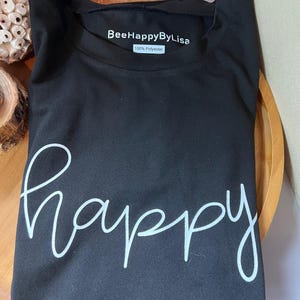 Peut inclure: T-shirt noir avec le mot "happy" écrit en cursive blanche. Le t-shirt porte une étiquette indiquant "BeeHappyByLisa 100% Polyester."