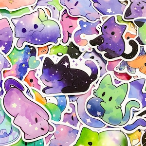 May include: A collection of colorful stickers featuring cute cartoon cats with galaxy-themed designs. The stickers are printed on white paper and have a glossy finish.
