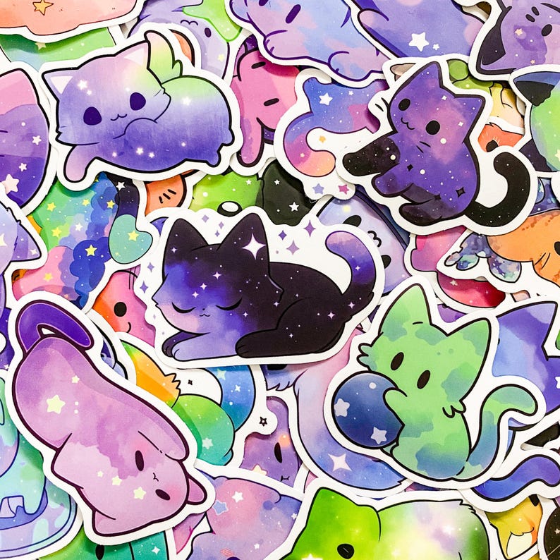 May include: A collection of colorful stickers featuring cute cartoon cats with galaxy-themed designs. The stickers are printed on white paper and have a glossy finish.