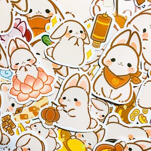 May include: A collection of white bunny stickers with various designs, including a bunny holding a lantern, a bunny sitting on a lotus flower, and a bunny with a red scarf. The stickers are on a white background.