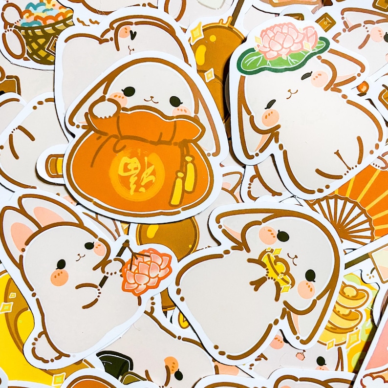 May include: A collection of stickers featuring cute cartoon bunnies in various poses. The bunnies are holding items like a red bag with Chinese characters, a lotus flower, and a gold coin. The stickers are on a white background.