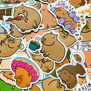 May include: A collection of whimsical stickers featuring cartoon capybaras in various poses. Some stickers show capybaras with a toothbrush, a cup of coffee, and a donut. The stickers are in shades of brown, pink, and blue.