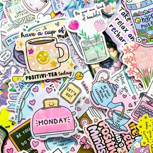 May include: A collection of colorful stickers with various designs and text. Stickers include a smiling mug with the text "POSITIVI-TEA today", a pink toaster with "MONDAY", and a teapot with "Be okay with not being everyone's".