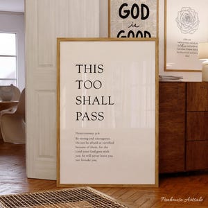 May include: A framed print with the words "THIS TOO SHALL PASS" in a minimalist style. Below the text is a Bible verse from Deuteronomy 31:6. The print is in a light-colored frame, with other framed art in the background.