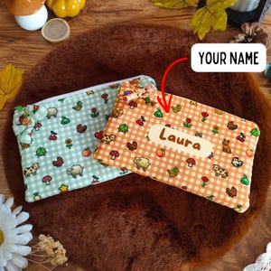 Op de afbeelding: Twee rechthoekige stoffen etuis met een gingham-patroon en kleine pixel-art iconen van boerderijdieren, fruit en paddenstoelen. De ene etui is lichtgroen, de andere is oranje met een wit label waarop de naam Laura staat weergegeven.