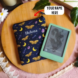 Puede incluir: Una funda de tela azul oscuro con un estampado de lunas crecientes amarillas, polillas marrones, cristales morados y ramas de lavanda, con el nombre Helena impreso en blanco. Al lado, un lector electrónico verde claro muestra una portada.