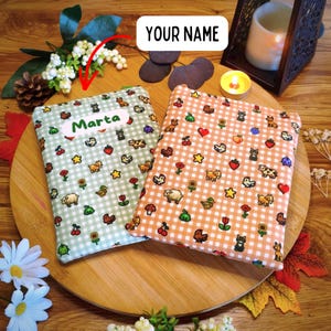 Può includere: Due astucci rettangolari in tessuto con motivo a quadretti vichy in verde chiaro e arancione. Gli astucci sono decorati con piccole icone in stile pixel-art di animali, frutta e fiori. Un astuccio presenta l'etichetta con il nome Marta.