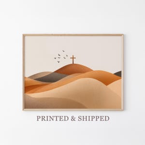 May include: A framed print featuring a minimalist landscape with rolling hills in shades of brown and beige. A wooden cross sits atop the highest hill, with a flock of birds flying above. The artwork has a serene and spiritual theme.