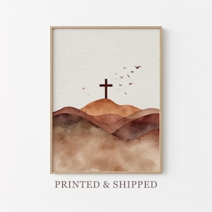 May include: A framed watercolor print featuring a brown cross atop rolling hills. The artwork uses shades of brown and tan, with birds flying in the sky. The words "PRINTED & SHIPPED" are printed at the bottom of the frame.
