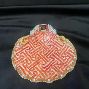 May include: A white seashell dish with a red and orange geometric pattern. The dish is trimmed with gold paint and rhinestones. A red jewel is set in the center of the dish.