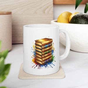 May include: White ceramic mug featuring a stack of golden books with colorful spines and a watercolor splash design. The books have blue, green, red, and purple spines. The mug is on a beige coaster.