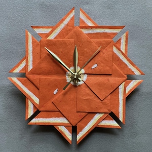 May include: Orange and white paper clock with gold hands. The clock is made of folded paper and has a starburst design.