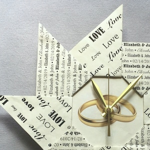 May include: A white origami clock with gold hands and two gold rings in the center. The clock is decorated with the words "Love" and "Elizabeth & John" repeated multiple times.