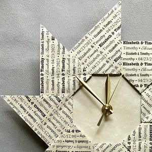 May include: A beige paper origami star clock with gold-colored hands. The star is decorated with the text "Elizabeth & Timothy" and various dates.
