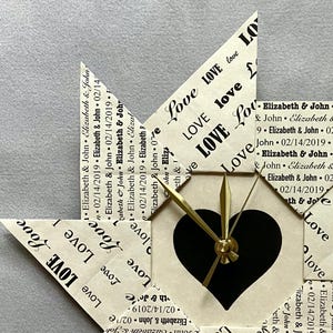 May include: A white origami clock with a black heart-shaped face and gold hands. The clock is decorated with the word "love" repeated many times and the names "Elizabeth & John" and the date "02/14/2019".