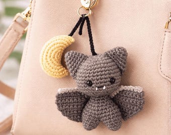 May include: A handmade crochet bat and crescent moon keychain in gray and yellow, hanging from a beige handbag. The bat has small wings and a cute face. The keychain is attached to a gold-colored clasp.