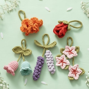 May include: A collection of handmade crochet flowers in various colors, including orange, red, pink, and purple. Each flower has a loop for hanging. The flowers are arranged on a light green surface with white flower accents.