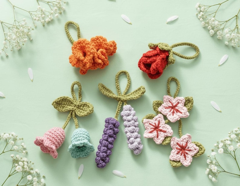 May include: A collection of handmade crochet flowers in various colors, including orange, red, pink, and purple. Each flower has a loop for hanging. The flowers are arranged on a light green surface with white flower accents.