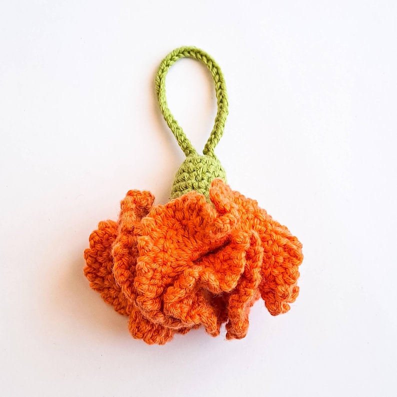 May include: A handmade crochet flower in vibrant orange, featuring detailed petal work. The flower has a green stem and a loop for hanging. The craftwork is set against a plain white background, highlighting the texture and color of the yarn.