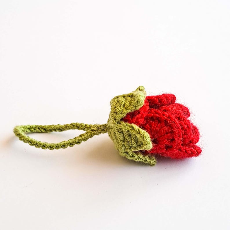 May include: A handmade crochet rose in vibrant red, with green leaves and a stem loop. The rose is detailed with textured stitches, showcasing a craft-like aesthetic. The loop stem adds a functional or decorative element.