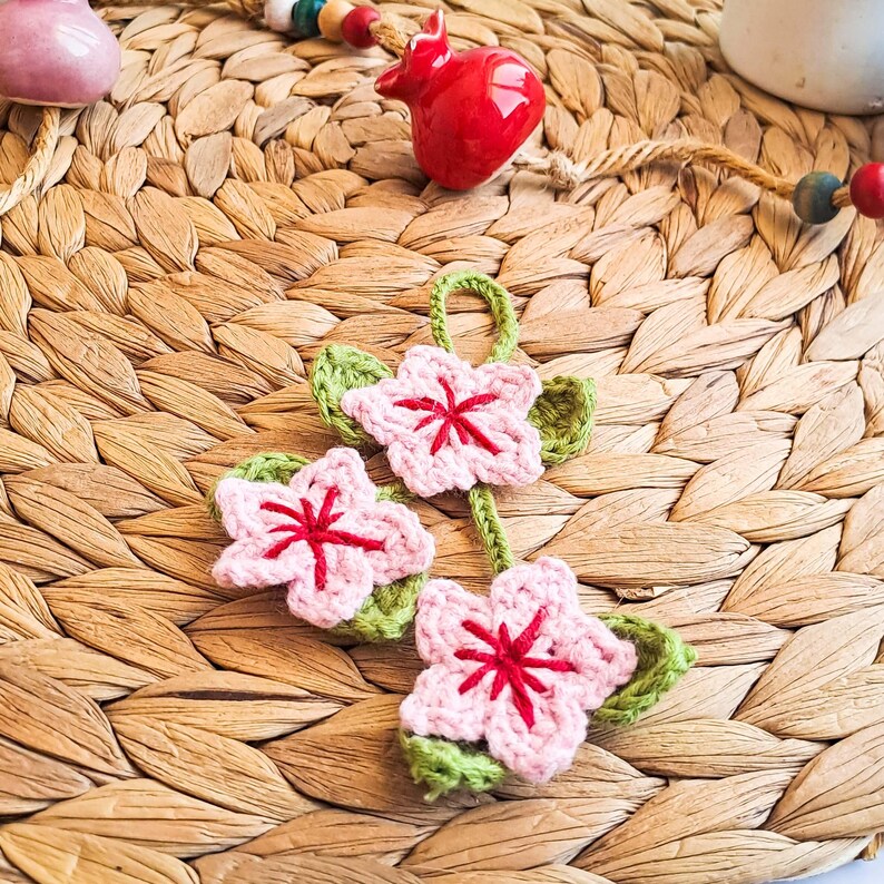 May include: Three handmade crochet flower ornaments. Each flower is pink with a red center and green leaves. The flowers are connected by green crochet stems. The background is a woven, light brown mat.