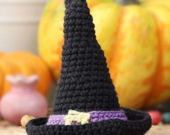 May include: A black crocheted witch's hat with a purple and yellow band. The hat is sitting on a wooden surface with pumpkins in the background.
