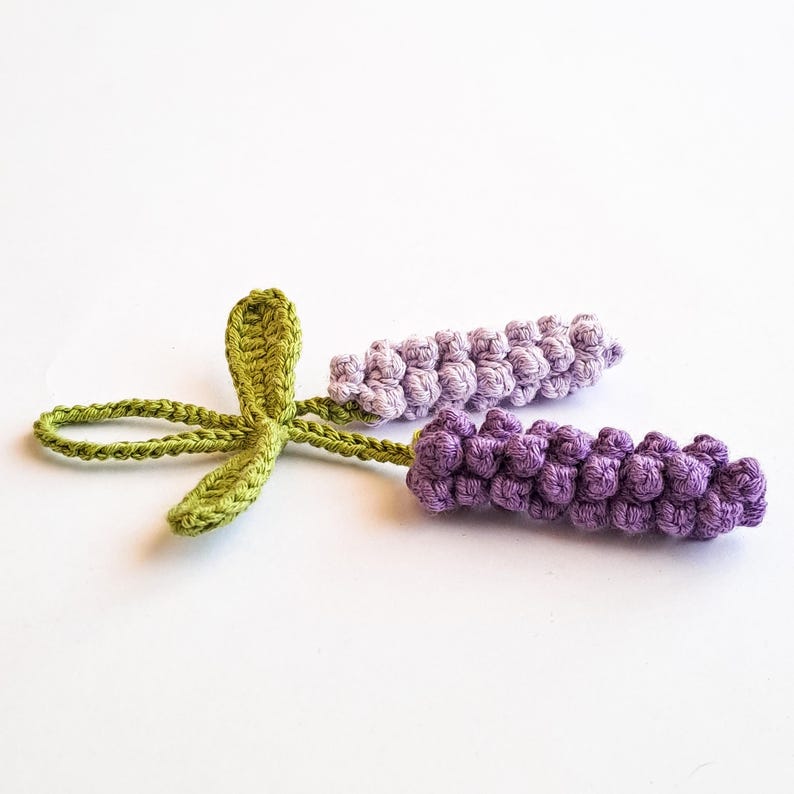 May include: Crocheted lavender sprigs in shades of purple and green. The lavender buds are a deep purple, while the leaves and stems are a vibrant green. The handmade craft is set against a white background.