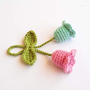 May include: Crocheted floral embellishment featuring a green leaf cluster and two flowers. One flower is pink, the other is light green. The stems and leaves are a vibrant green color, and the flowers have a textured appearance.