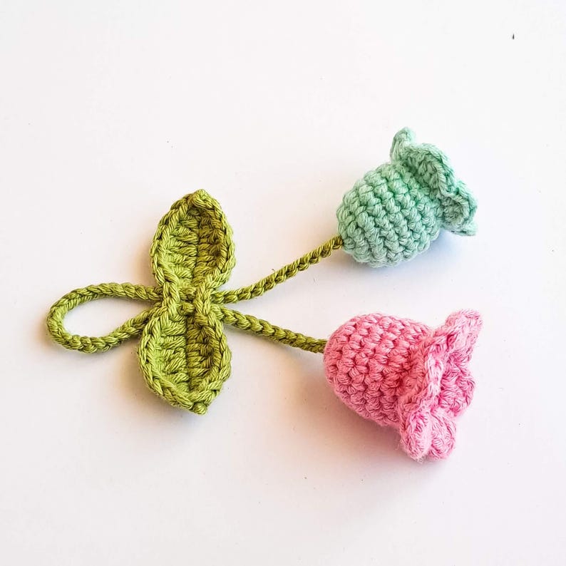 May include: Crocheted floral embellishment featuring a green leaf cluster and two flowers. One flower is pink, the other is light green. The stems and leaves are a vibrant green color, and the flowers have a textured appearance.
