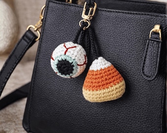 May include: Crocheted Halloween-themed bag charms. One charm is a white eyeball with red veins and a black pupil. The other is a candy corn charm with orange, yellow, and white sections. Both charms are attached to a black purse.