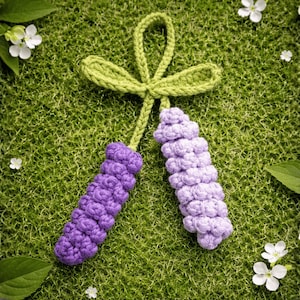 May include: A handmade crochet lavender sprig ornament with a green stem and bow. The lavender is a deep purple color, and the bow is a light green. The ornament is set against a green background with white flowers and leaves.