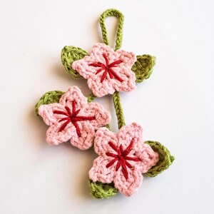 Handmade crochet cherry blossom keychain featuring three light pink flowers with red centers and green leaves.
