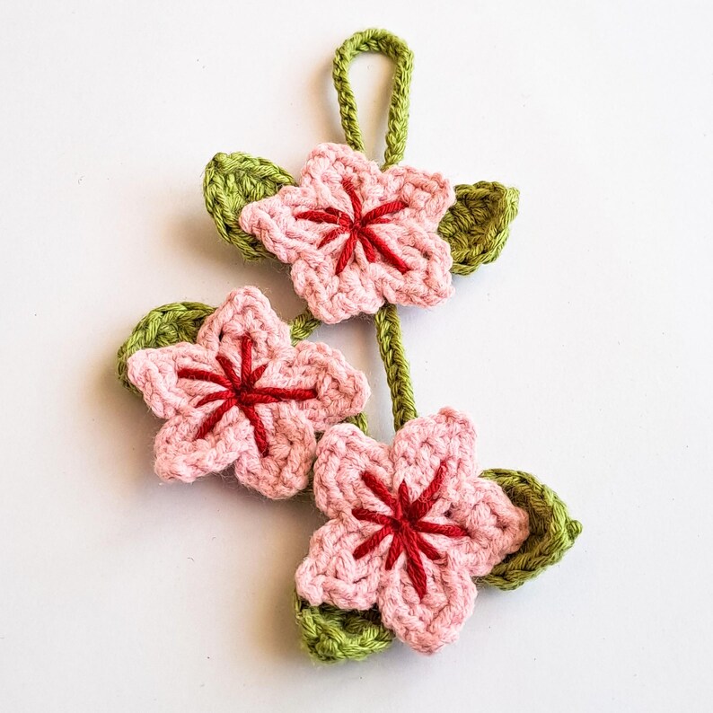 Handmade crochet cherry blossom keychain featuring three light pink flowers with red centers and green leaves.