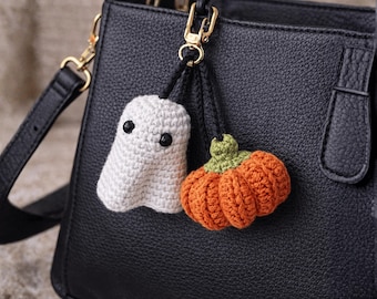 May include: Crocheted Halloween-themed keychains. One keychain is a white ghost with black eyes, and the other is an orange pumpkin with a green stem. Both are attached to a black leather handbag with gold hardware.